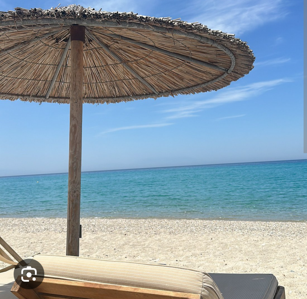 a beach chair and umbrella in the foreground, crystal clear greek water behind with blue skies