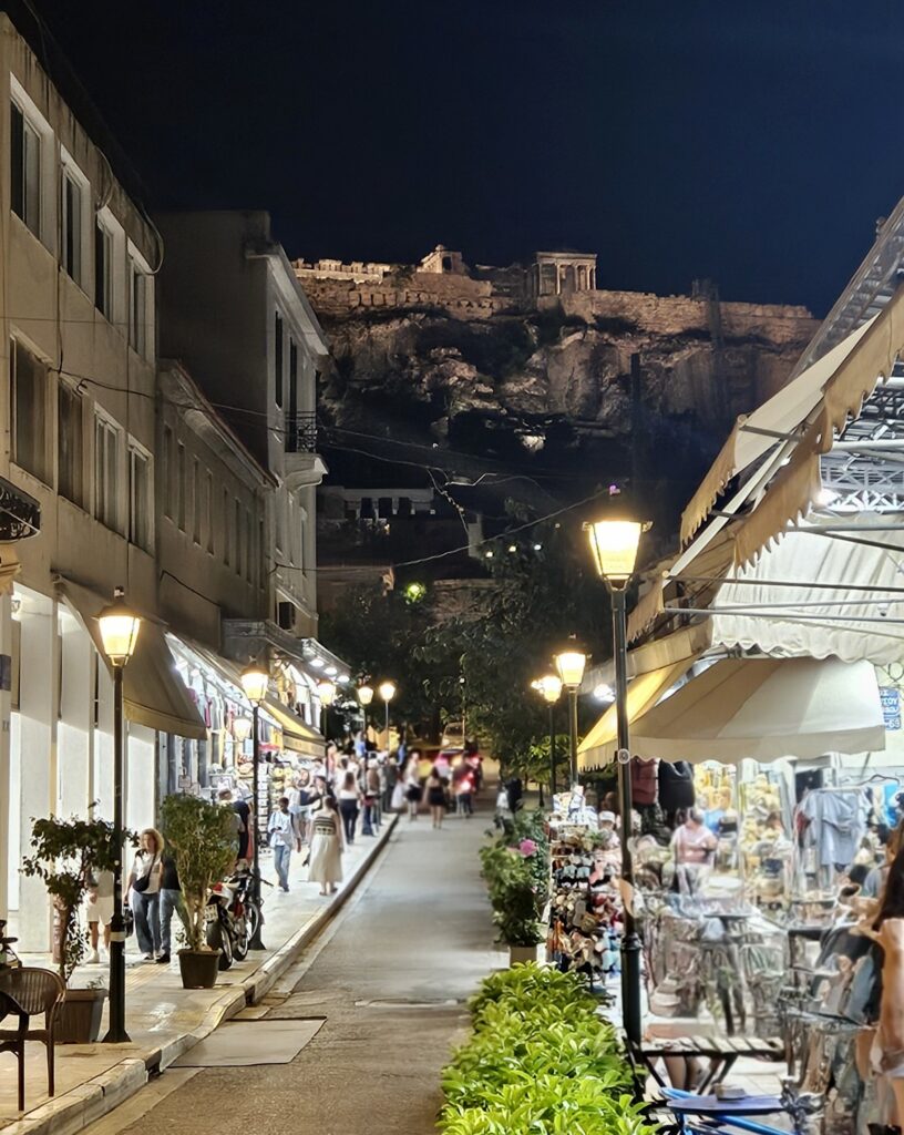 an evening shot of a street in the old town of plaka athens with shops on the left and right, the acropoplis overhead