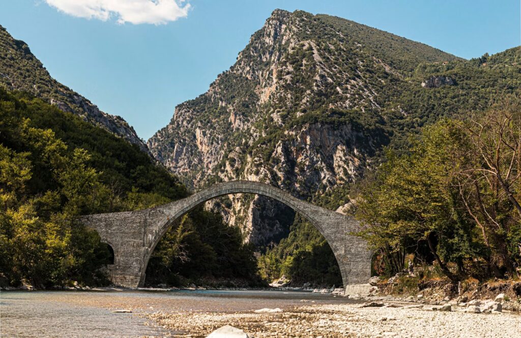 an old arch bridge over a shallow, crystal clear riverbed in Arta, Greece. Mountains behind and blue skies.