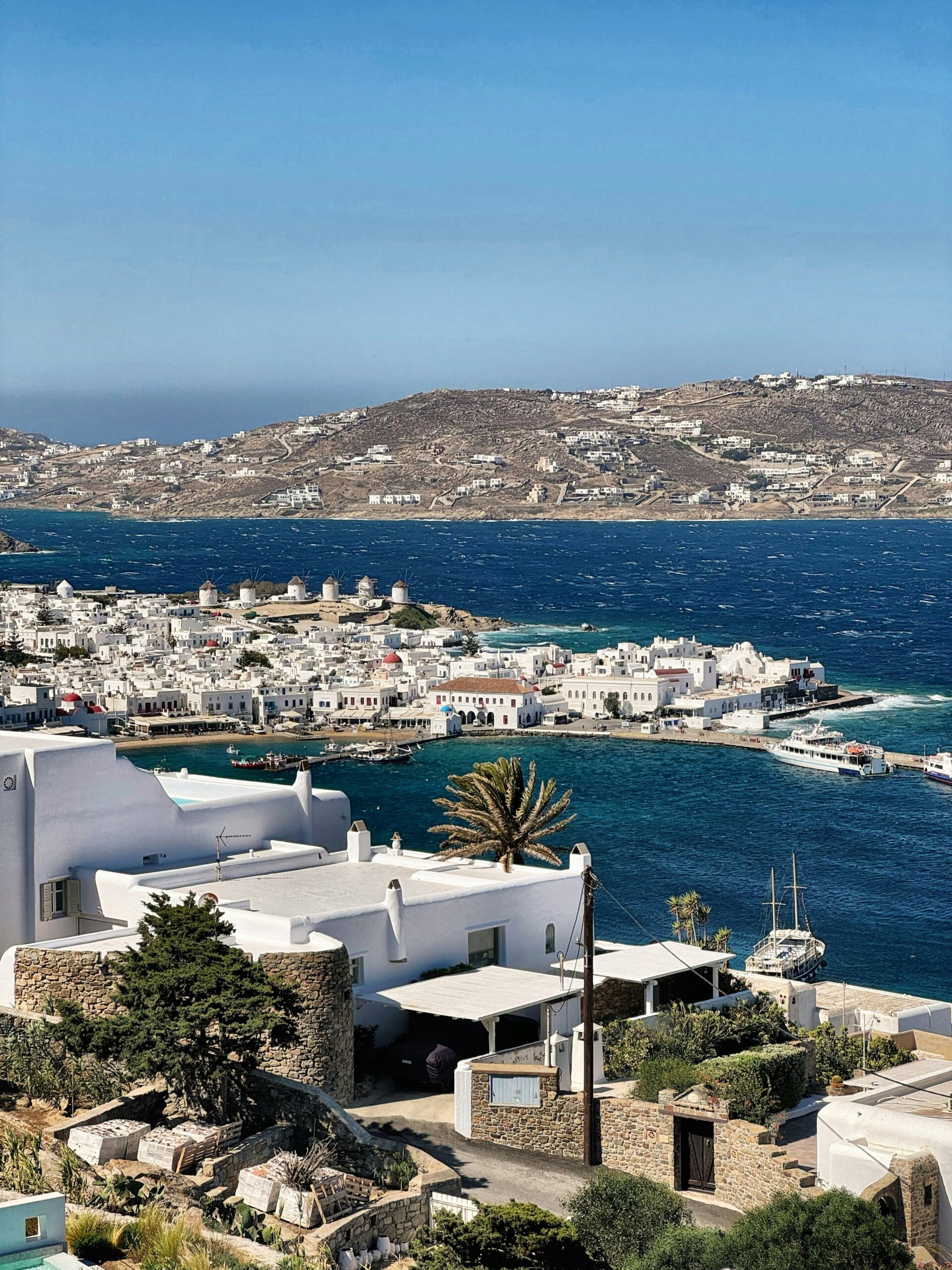 a photo of mykonos town and port from above, blue waters and moutains in the background