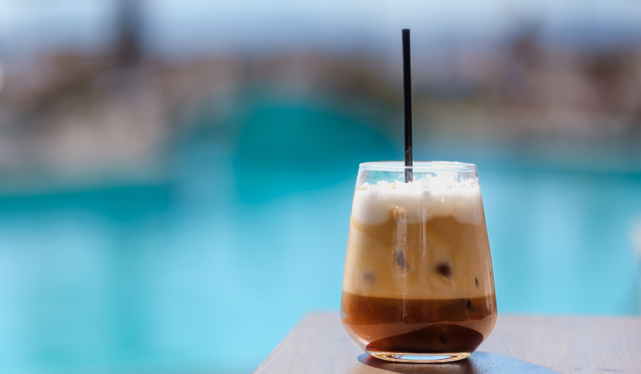 a freddo cappuccino with dark brown at the bottom, then caramel colored foam topped with whipped white cream. azure water blurred in the background