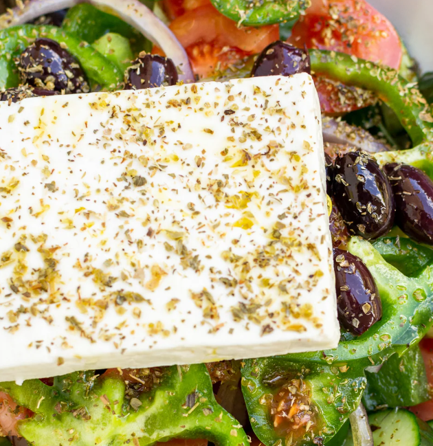 thick slice of feta with oregano and olive oil over a greek salad with tomato green pepper and olives